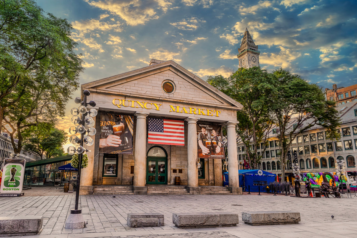 Quincy Market in Boston illuminated by warm morning light beneath dramatic clouds, with an American flag hanging over the entrance and the Custom House Tower rising in the background.