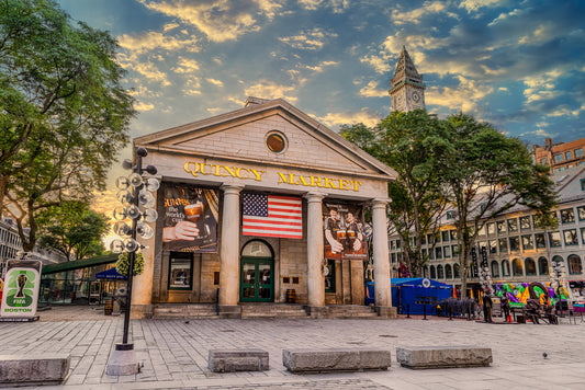 Quincy Market in Boston illuminated by warm morning light beneath dramatic clouds, with an American flag hanging over the entrance and the Custom House Tower rising in the background.