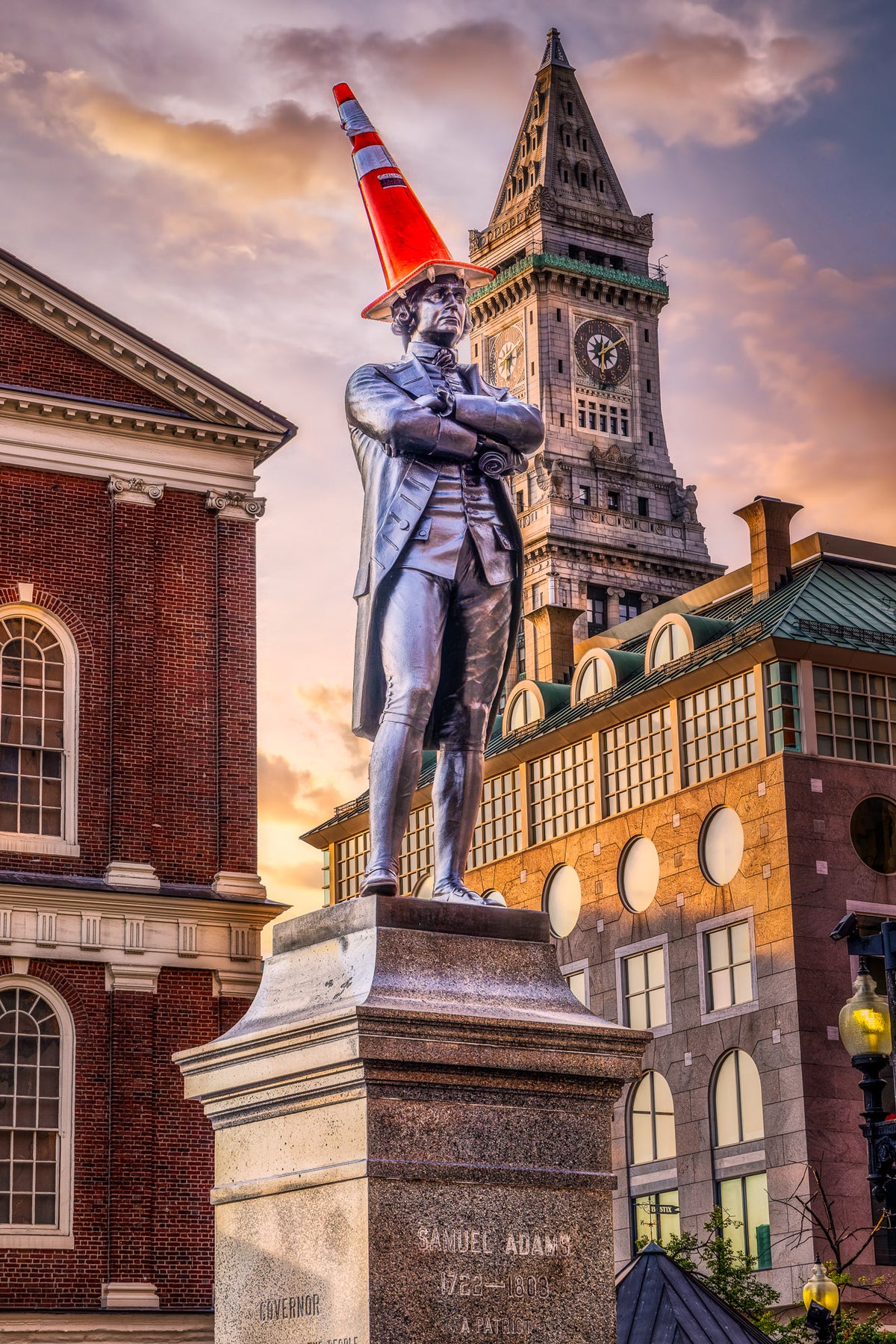 Samuel Adams statue outside Faneuil Hall in Boston wearing an orange traffic cone at sunrise, with the Custom House Tower rising behind him.