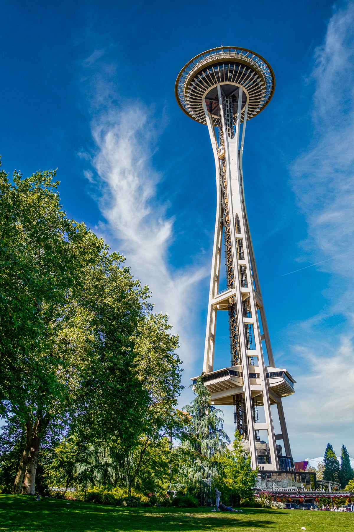 Seattle's Space Needle towering above green trees at Seattle Center beneath a vivid blue sky filled with sweeping white clouds.