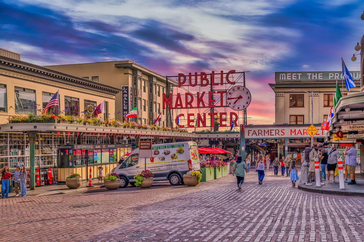 Pike Place Market's iconic Public Market Center neon sign and clock above busy brick streets in downtown Seattle beneath a colorful pink and blue sunset.